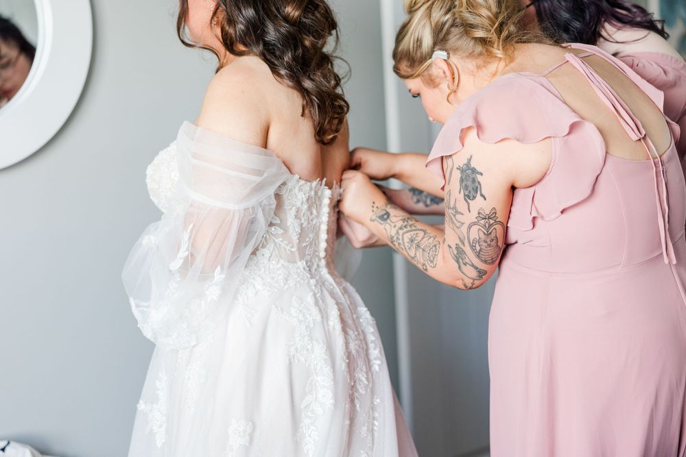 Bridesmaid in pink dress helping bride lace up her white wedding gown before the ceremony.