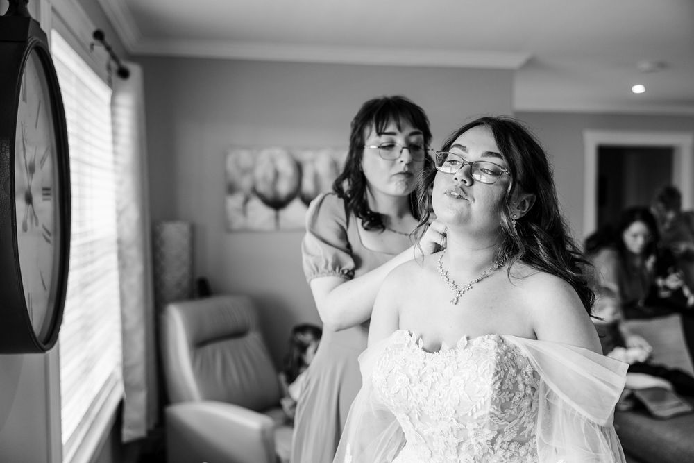 Bridesmaid helping bride with necklace while getting ready in a candid black and white wedding preparation photo.