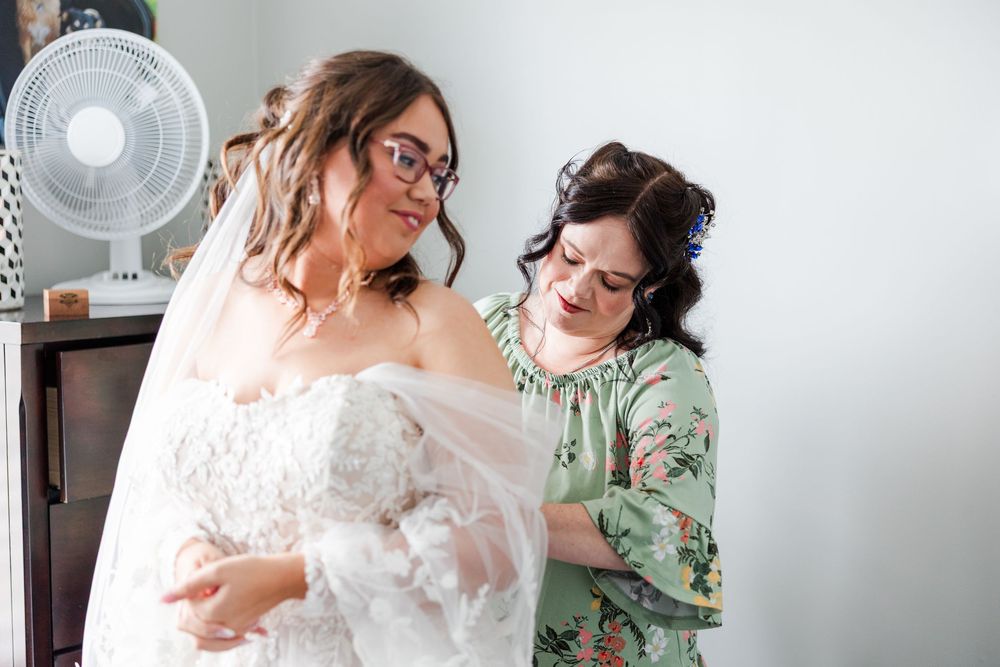 Bride in lace wedding dress getting help from bridesmaid adjusting veil in bright white dressing room.