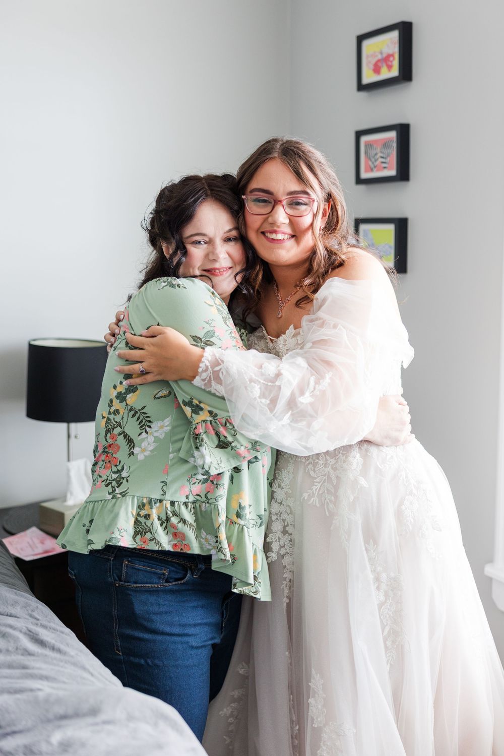 A bride in a white lace gown hugs a woman in a floral green top, sharing a joyful moment before the wedding ceremony.
