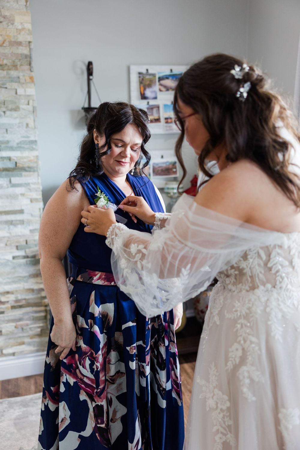 Bride in white lace gown pins boutonniere on woman in blue floral dress during wedding preparations.