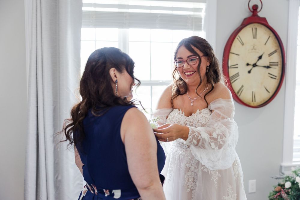 Bride in lace gown laughing with bridesmaid in navy dress while getting ready near a vintage red wall clock.