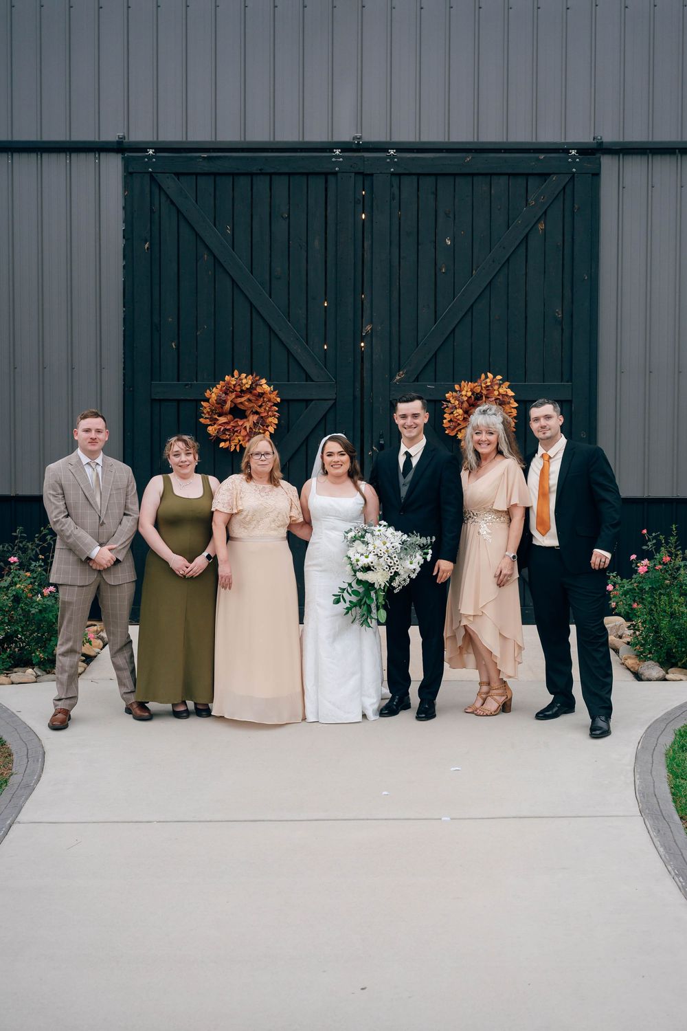 Wedding party poses before dark barn doors, bride in white gown holding bouquet, flanked by family in formal attire.