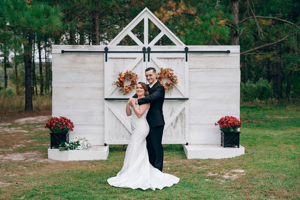 Bride and groom embrace in front of a rustic white barn door backdrop with fall floral wreaths and mums outdoors.