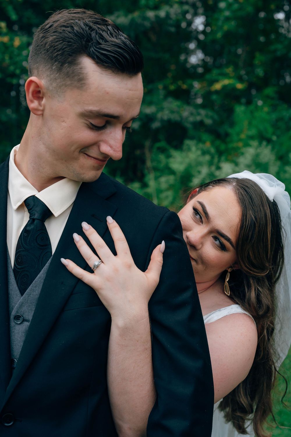 A bride in a white veil rests her head on a groom in a black suit, showcasing her engagement ring outdoors.