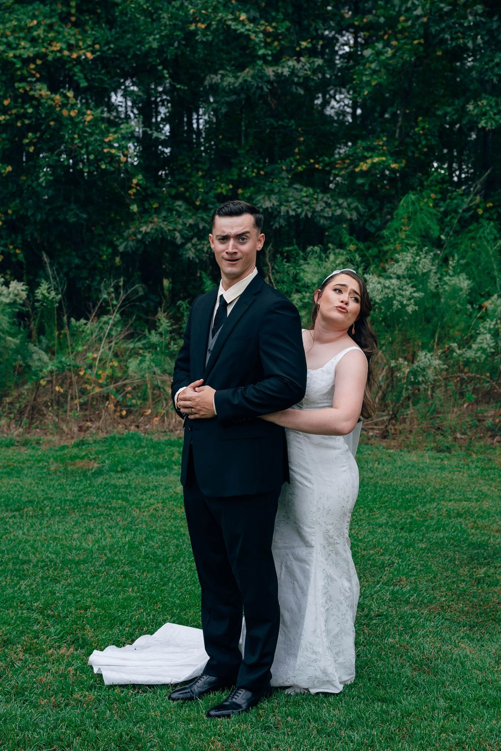 Bride in white gown embraces groom in black suit outdoors on green lawn with lush forest backdrop.