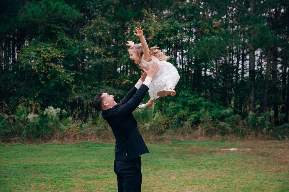 Man in black suit joyfully tosses young girl in white dress into the air outdoors near lush green trees.