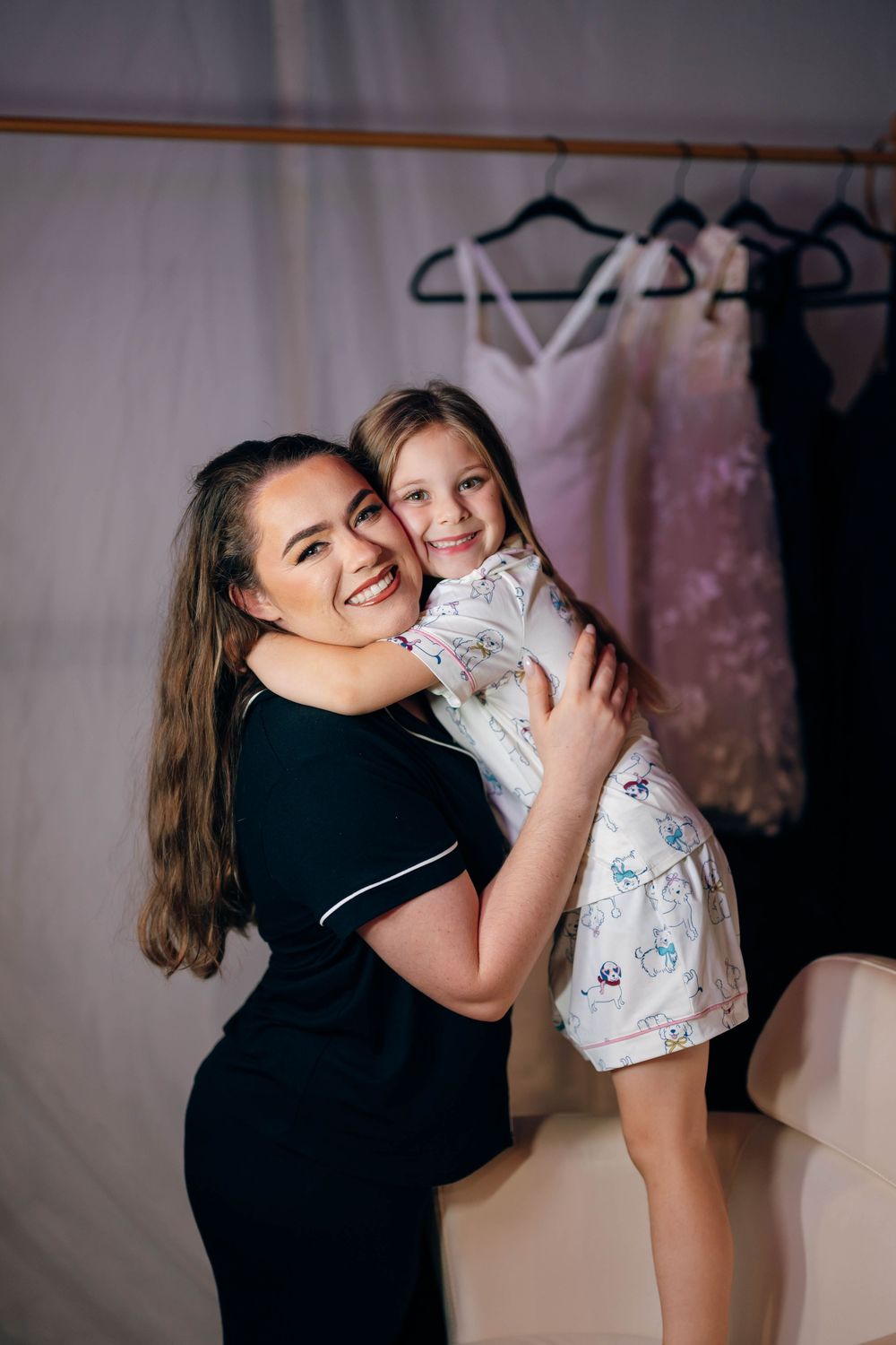 A woman in a black dress hugs a smiling young girl in a floral outfit, with dresses hanging in the background.