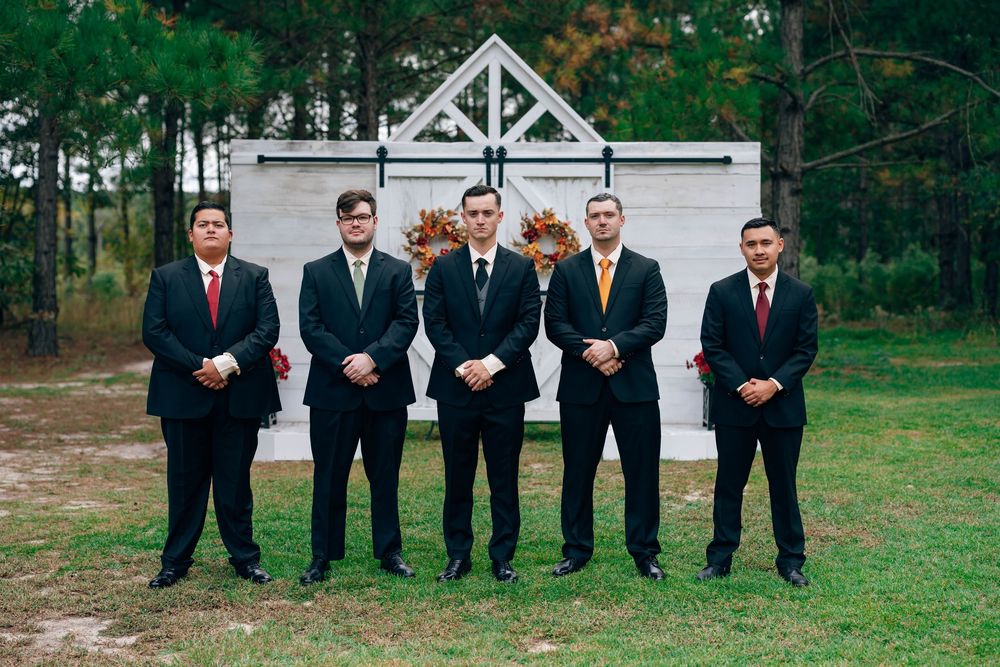 Six groomsmen in black suits with red and gold ties pose outdoors in front of a white barn door wedding backdrop.