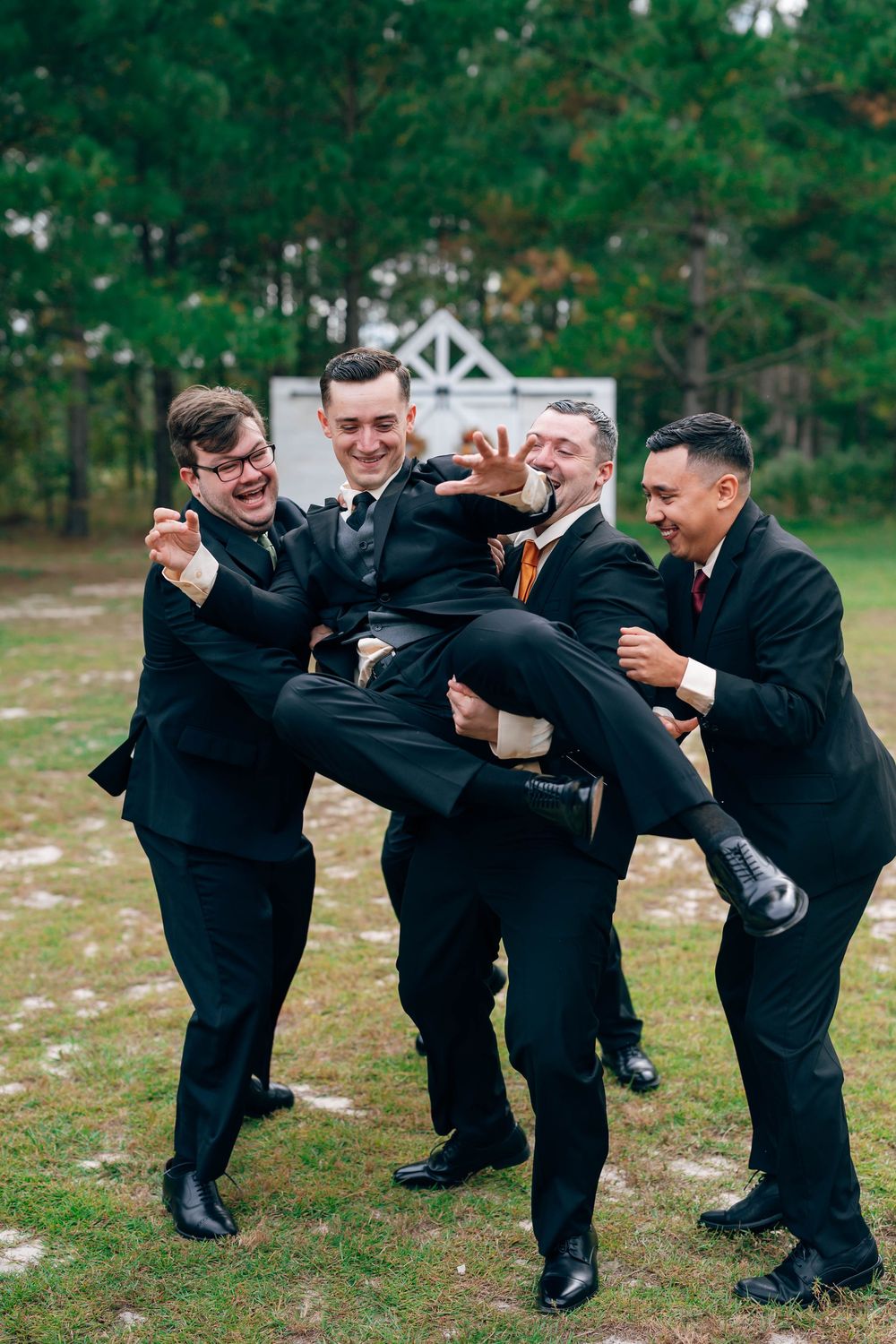 Groomsmen in black suits laughing and lifting the groom outdoors at a wedding celebration.