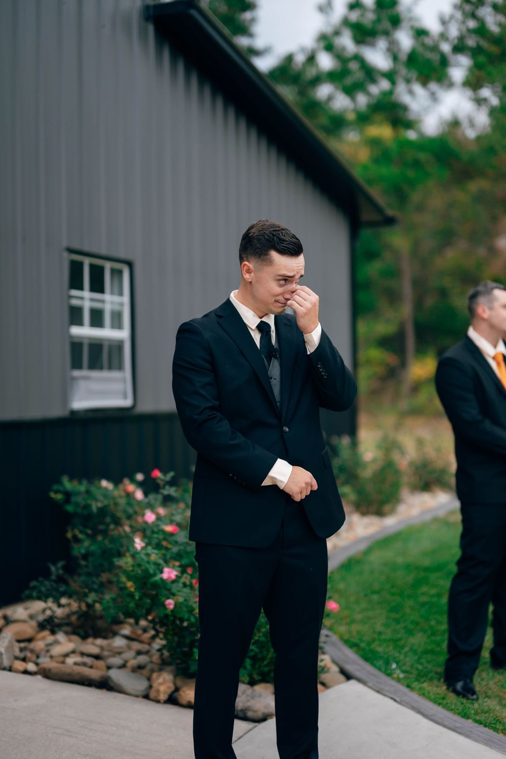 Emotional groom in black suit wiping tears outside barn venue on wedding day.