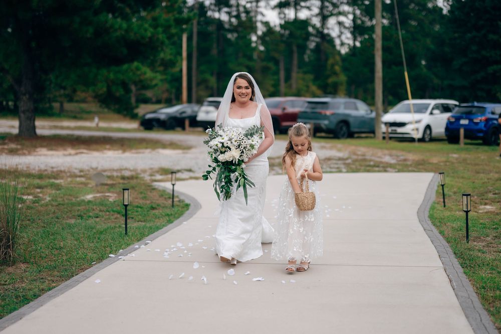 Bride in white gown and veil walks with flower girl down outdoor wedding aisle surrounded by pine trees.
