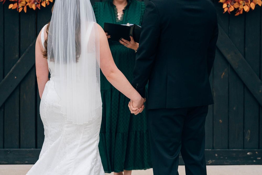 Bride and groom holding hands at outdoor wedding ceremony with officiant in green dress before dark barn doors.