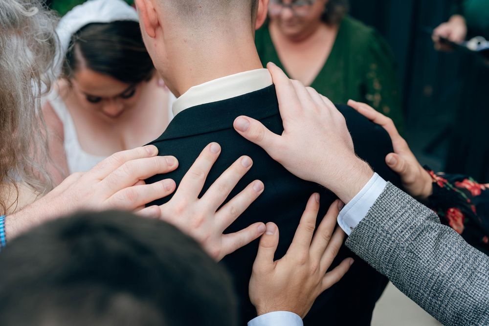 Multiple hands placed on the back of a groom in a black suit during a wedding blessing ceremony.