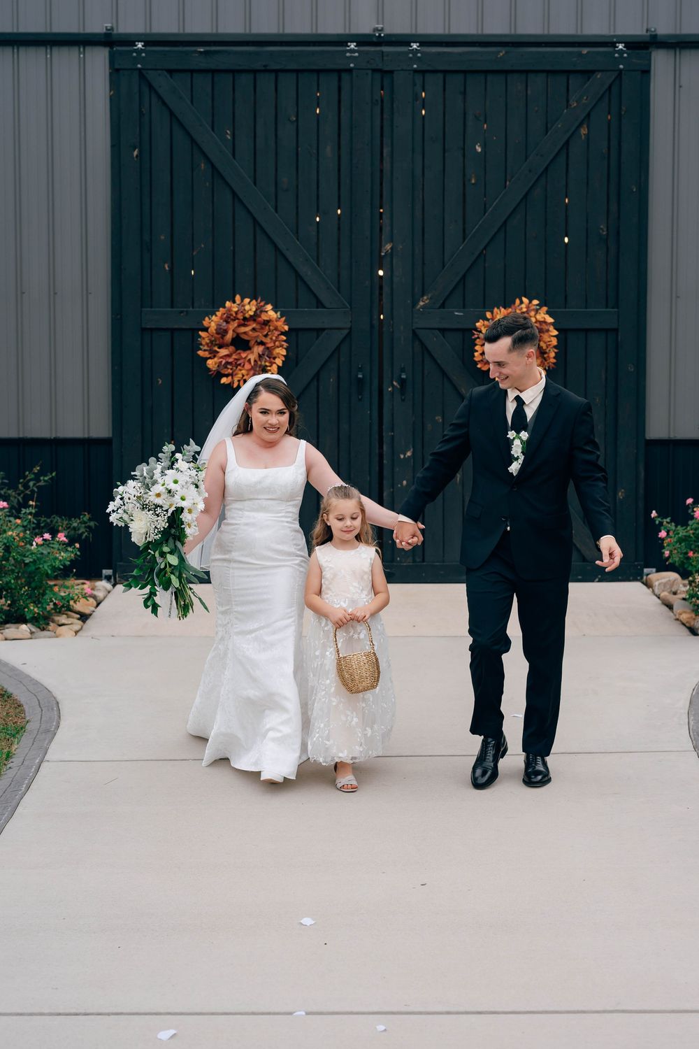 Bride and groom walk hand-in-hand with flower girl outside rustic barn venue decorated with fall wreaths.