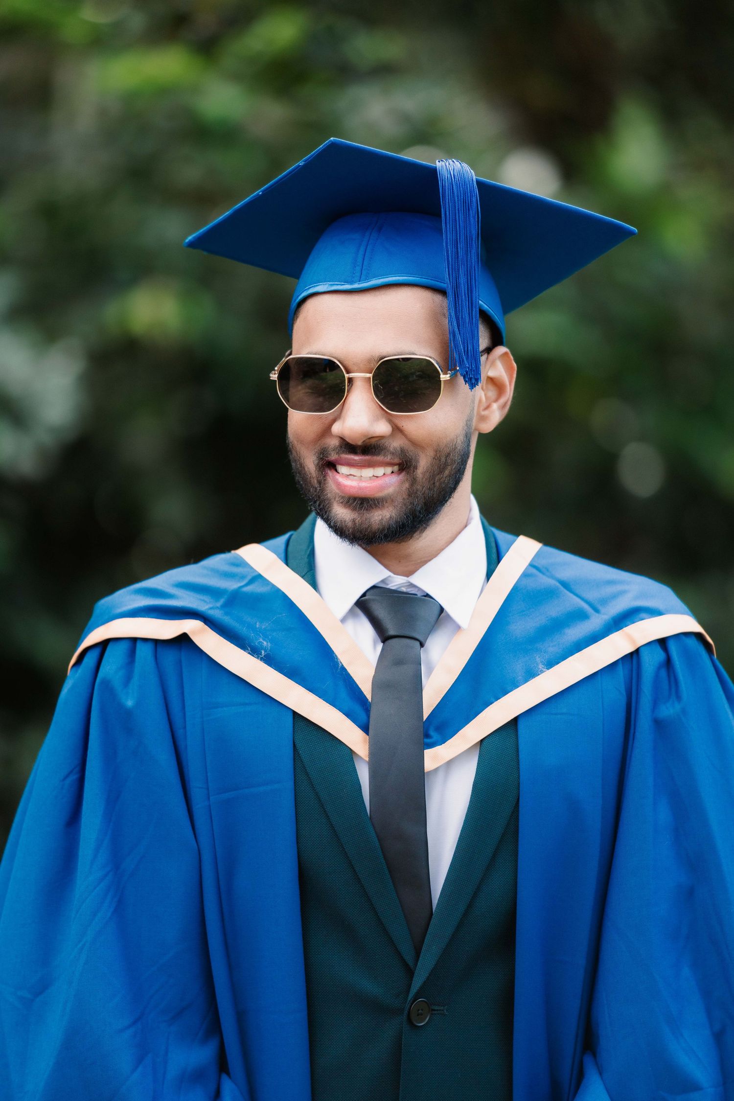 Smiling graduate in sunglasses, a blue cap and gown with gold trim, and a green jacket and tie, posing outdoors in front of green foliage.