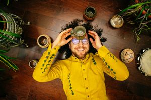 Smiling bearded man in a yellow leaf-print shirt lies on a wood floor holding a small cup over his eyes, surrounded by plants and drums.