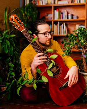Bearded man in glasses and yellow sweater sits among leafy houseplants holding a red acoustic guitar in front of a wooden bookshelf.
