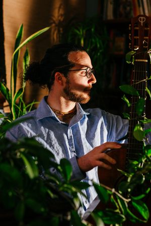 Bearded man with glasses and a loose blue shirt plays an acoustic guitar surrounded by green houseplants in warm sunlight.