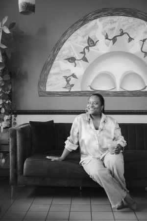 Smiling woman in a satin shirt and wide-leg pants sits on a tufted velvet sofa beneath a sun mural in a black and white photo.