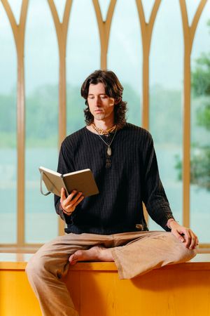 Man with wavy dark hair and layered necklaces sits barefoot on a yellow ledge, reading a book beside tall arched windows.