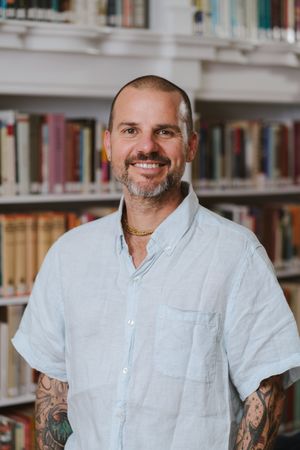 Smiling man with a goatee and tattooed forearms wears a light blue linen shirt while standing in front of library bookshelves.