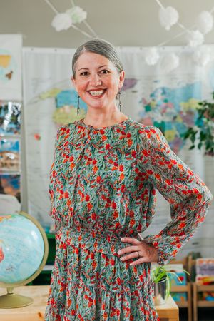 Smiling woman with gray hair in a floral dress stands with her hand on her hip in a bright classroom with a world map and globe.