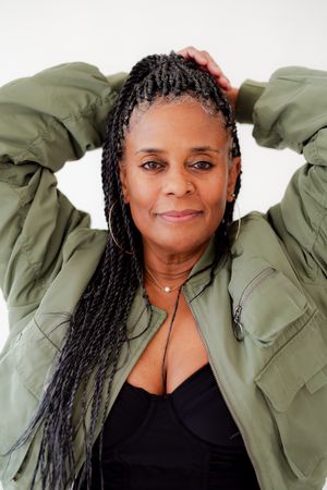 Woman with long gray-streaked braids poses with hands behind her head, wearing an olive bomber jacket and black top.