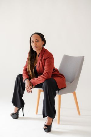 Woman with long braids in a rust blazer, black wide-leg pants and platform heels sits on a gray mid-century chair against a white backdrop.