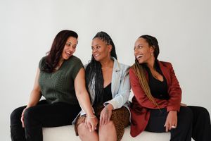 Three women laugh and share a joyful moment while sitting close together on a white bench against a bright, neutral studio backdrop.