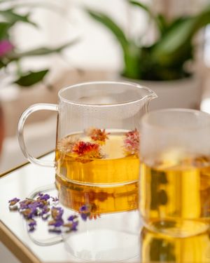 Glass teapot filled with golden flower tea and floating dried blossoms beside a glass cup and scattered purple petals on a mirrored tray.