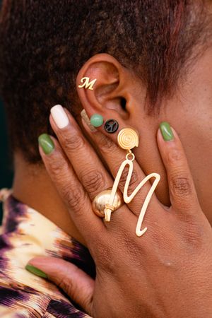 Close-up of a hand touching an ear adorned with gold initial M jewelry, a spiral earring, and green nail polish.