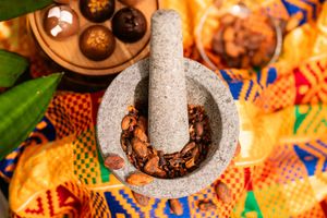 Stone mortar and pestle filled with roasted cacao beans on a colorful woven textile, beside a wooden tray of chocolate truffles.