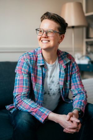 Smiling person with short hair and glasses wearing a plaid shirt over a graphic tee, seated on a couch with hands clasped.