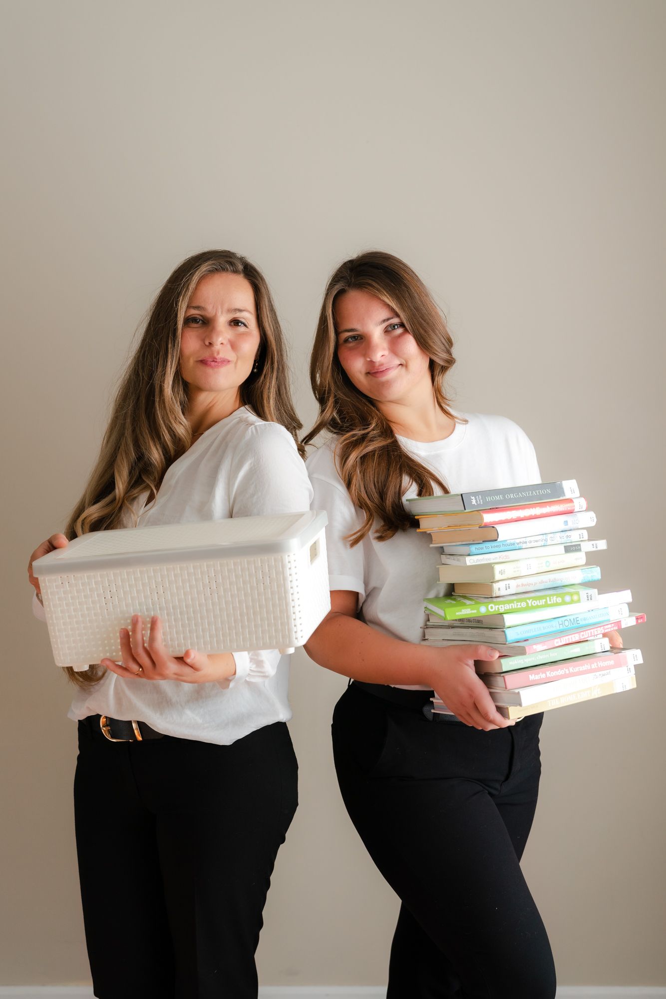 Two smiling women in white tops and black pants stand back to back, one holding a white woven storage basket and the other a stack of books.