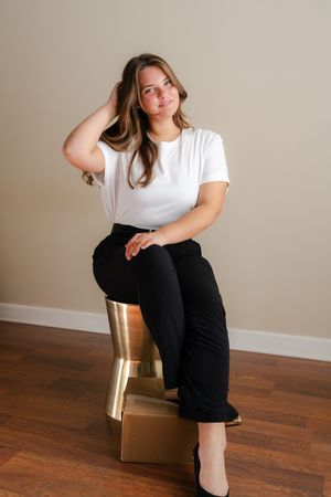 Woman in a white t-shirt and black pants sits on a gold stool, smiling with her hand in her hair, against a neutral wall and wood floor.