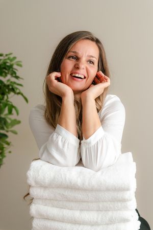 Smiling woman with long brown hair in a white blouse rests her chin on her hands atop a stack of folded white towels.