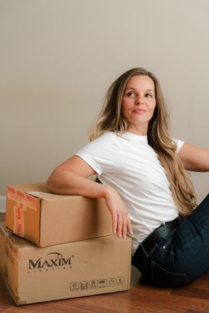 Woman with long hair in a white t-shirt and jeans sits on a wood floor, leaning on stacked Maxim Lighting cardboard boxes.