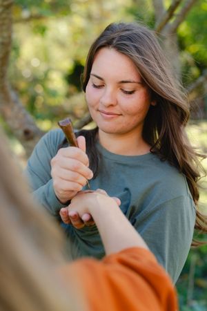 Young woman with long brown hair in a green shirt smiles while carefully using a small tool on another person's hand outdoors.