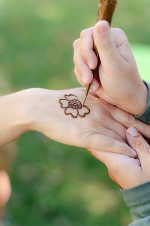 Artist painting a floral henna design on the back of a hand using a brush, with a blurred green outdoor background.
