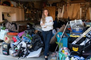 Woman with long hair stands with arms crossed in a cluttered garage overflowing with boxes, tools, bags and household items.
