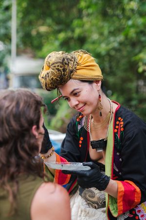 Smiling woman in a golden patterned head wrap and colorful embroidered jacket wearing black gloves while painting a customer outdoors.