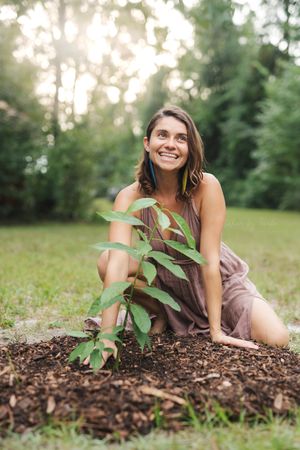 Smiling woman in a brown dress kneeling beside a young sapling planted in a mulched circle on a sunlit grassy lawn.