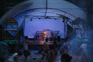 Musicians perform on an outdoor stage under a canopy at dusk while festival-goers dance in the foreground.