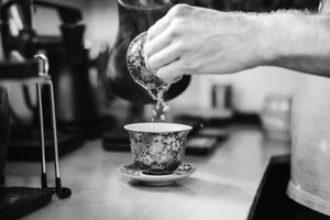 Black and white photo of a hand pouring tea from a small pot into an ornate floral porcelain teacup on a saucer.