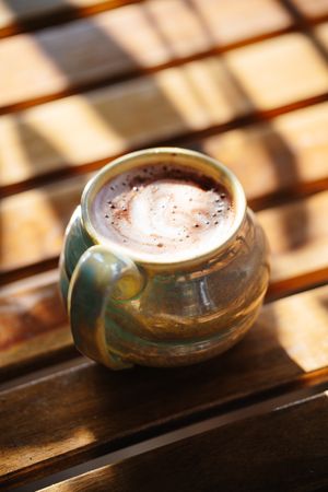 Handcrafted green and blue ceramic mug filled with frothy hot cocoa sitting on a sunlit rustic wooden slat table.