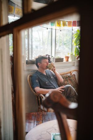 Man sipping a drink from a mug while seated in a wicker chair by a sunny window with potted plants and colorful prayer flags.