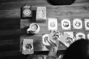 Woman arranging oracle cards in a row on a wooden table beside a steaming cup of tea and a card deck box in black and white.