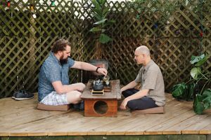 Two men sit on cushions at a low wooden table on an outdoor deck, sharing tea near a lattice fence and green plants.