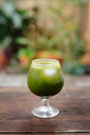 Fresh green juice with ice in a stemmed glass on a rustic wooden table, with blurred garden plants in the background.